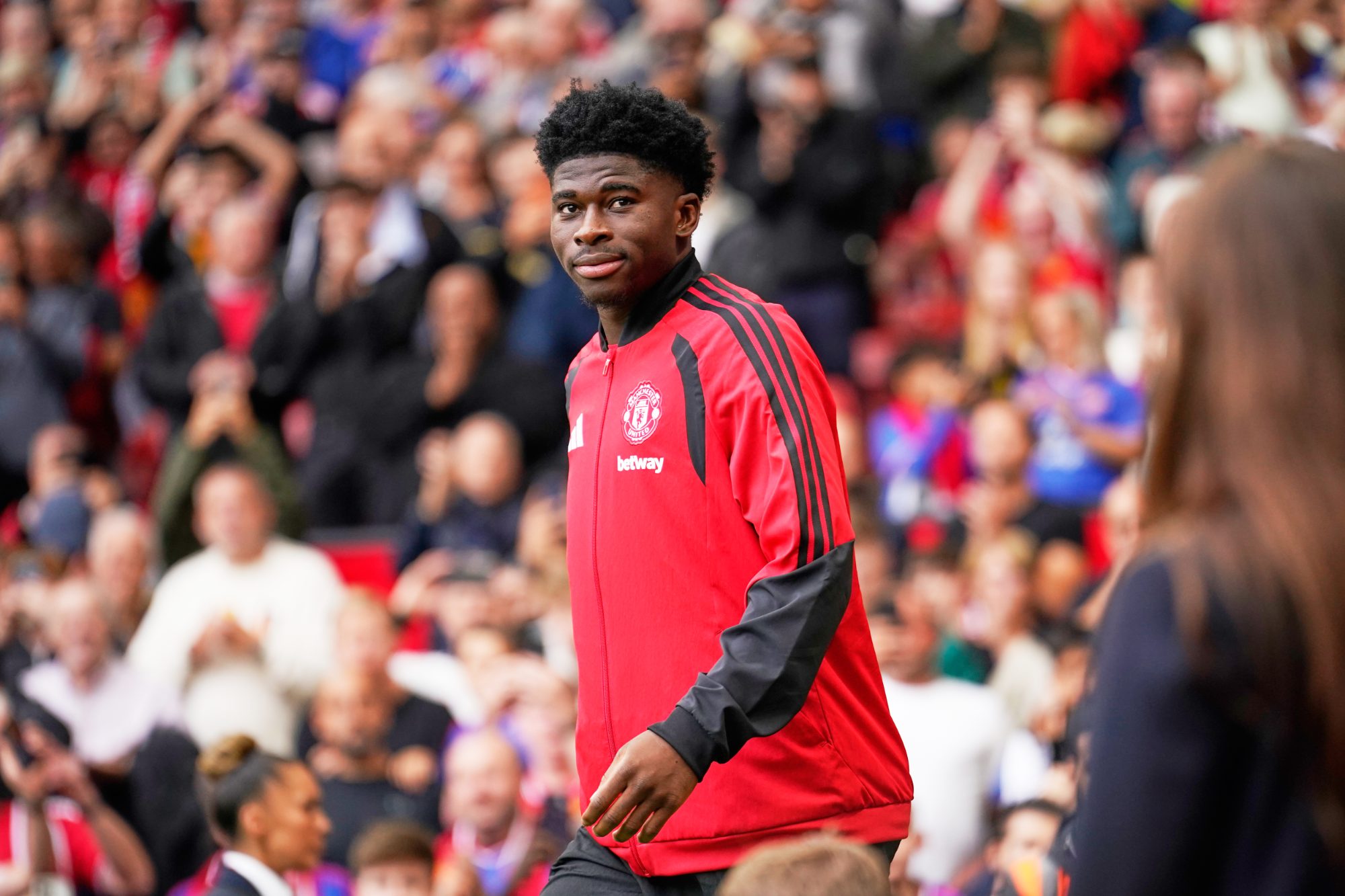 Manchester United player in a red training jacket walking on the pitch in front of a crowded stadium